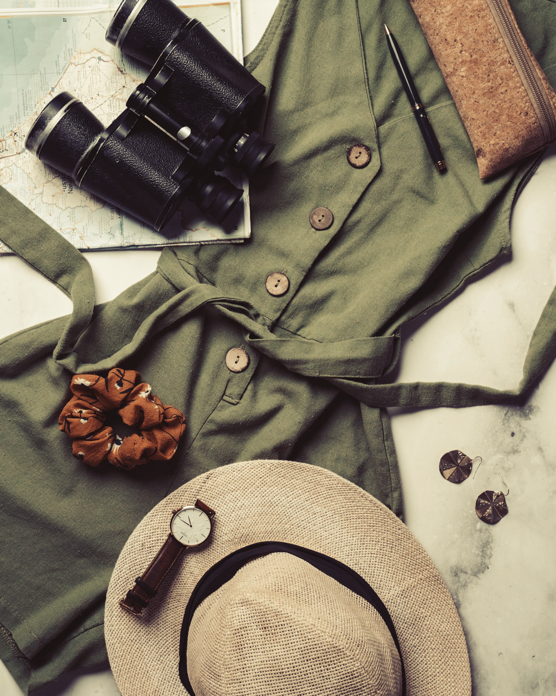 Green outfit, straw hat, black binoculars, and a small brown flower arranged on a light background.