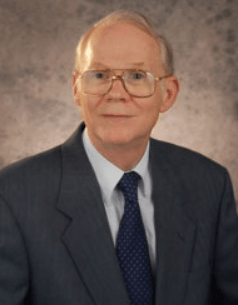 Portrait of a man in a dark suit, blue tie, and glasses against a mottled gray studio backdrop