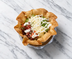 A crispy tortilla bowl with beans, cheese, and diced toppings on a marble surface.