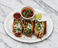 Three tacos with salsa and lime wedges on a white plate over a marble table.