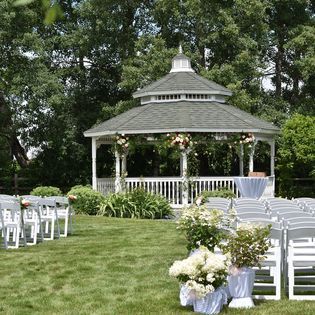 White wedding chairs set on a lawn before a decorated gazebo in a green outdoor garden.