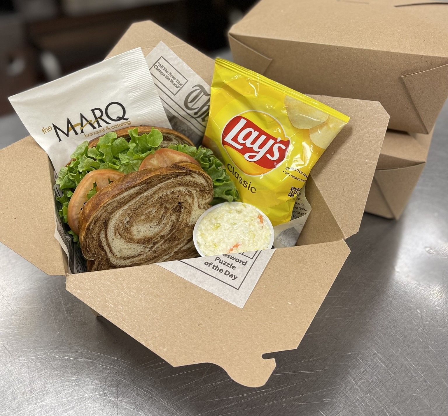 Open cardboard takeout box with a sandwich, Lay's chips, and a small side container on a table.