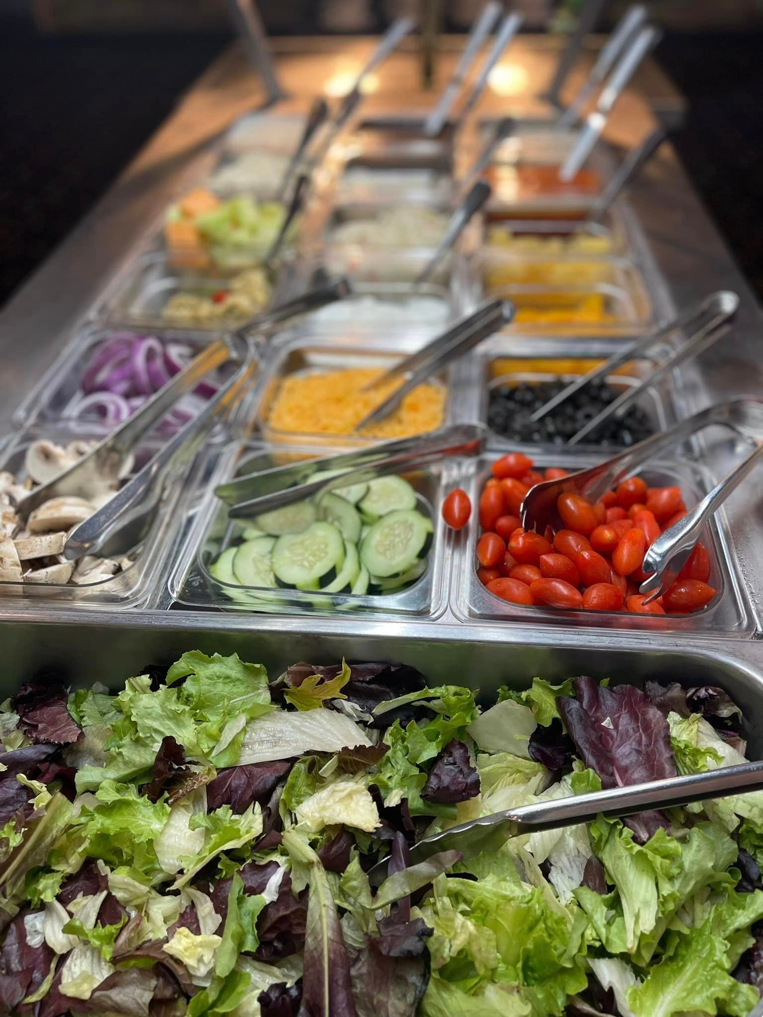 Salad bar with metal trays of toppings and leafy greens, including cucumbers and cherry tomatoes