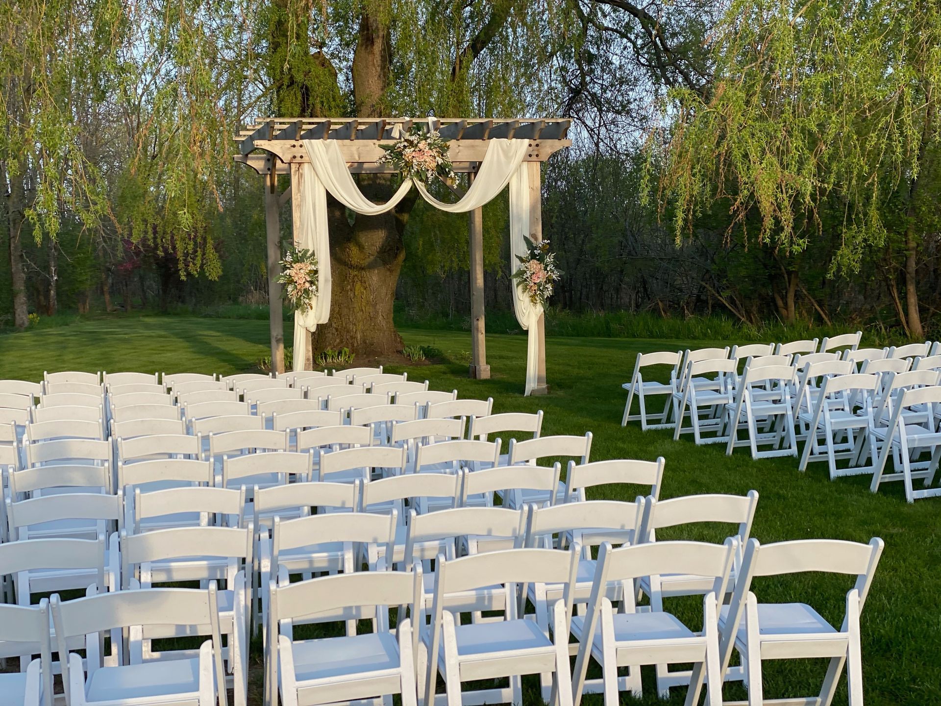 Outdoor wedding ceremony setup with white chairs and draped arch on a grassy lawn