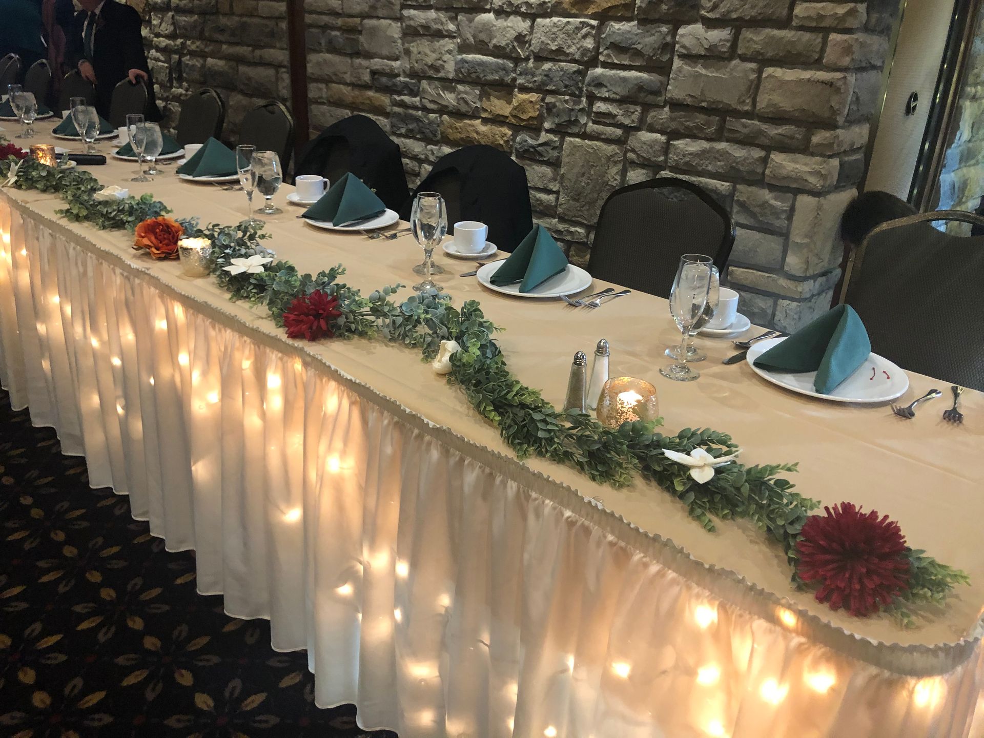 Decorated banquet table with fairy lights, greenery, and folded napkins against a stone wall