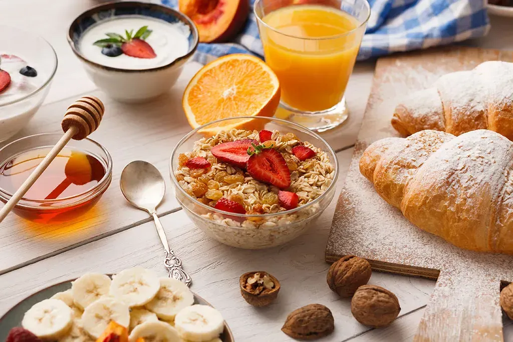 Breakfast spread with yogurt, fruit, orange juice, croissant, banana slices, and nuts on a table