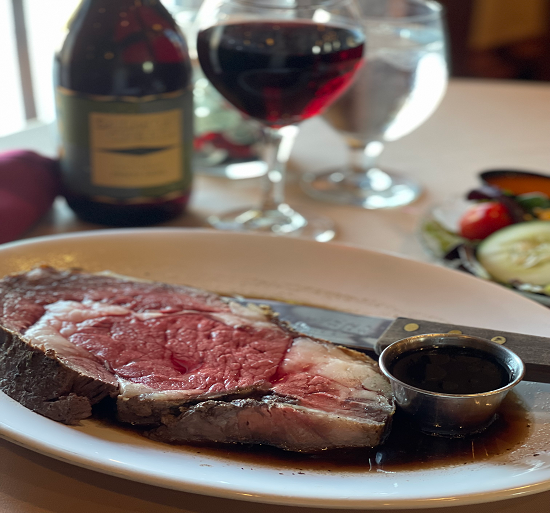 Steak on a white plate with red wine and sauces on a dining table, with a wine glass and bottle in the background
