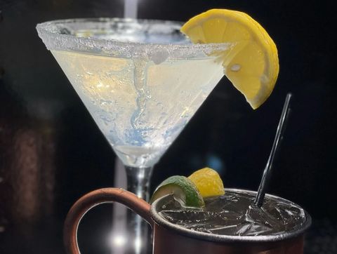 Two cocktails on a dark bar, one clear martini with a lemon twist and one dark drink with lime and straw