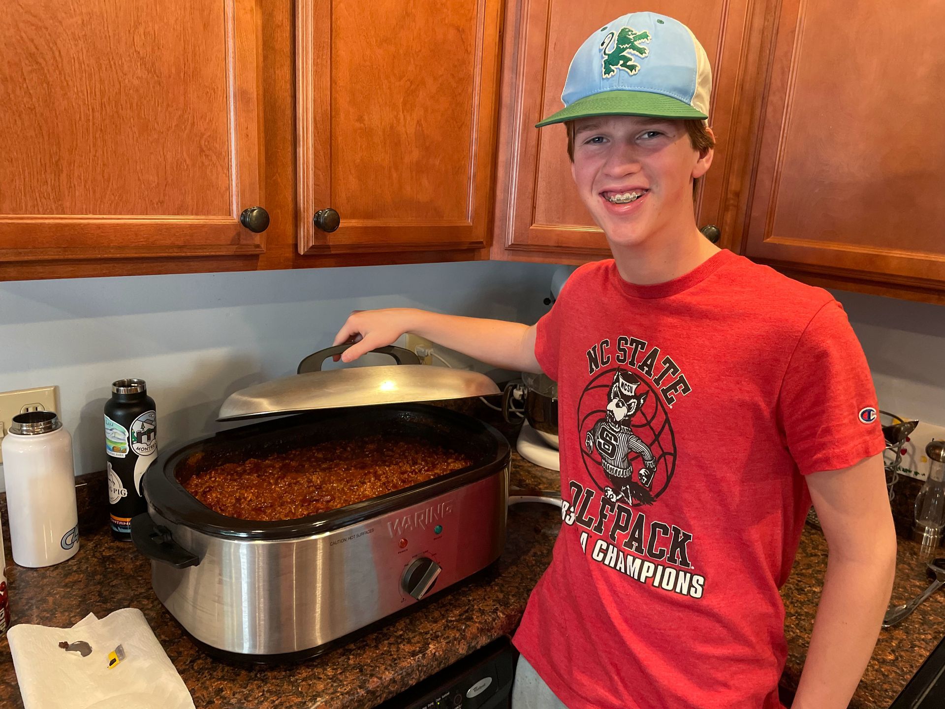 A boy in a red shirt is standing in front of a crock pot filled with chili.