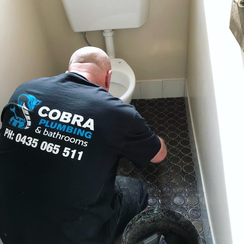 Plumber Working on a Toilet in a Small Bathroom. The Man Wears a Black Shirt With Business Logo — Cobra Plumbing & Bathrooms in Kiama, NSW