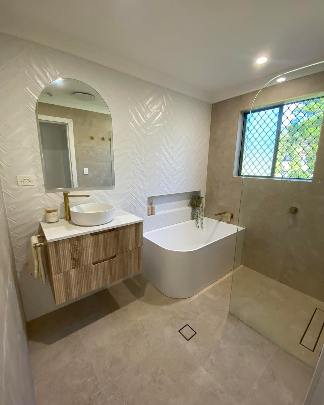 Modern Bathroom With a Wood Vanity, Arch Mirror, and a White Bathtub Next to a Window — Cobra Plumbing & Bathrooms in Kiama, NSW