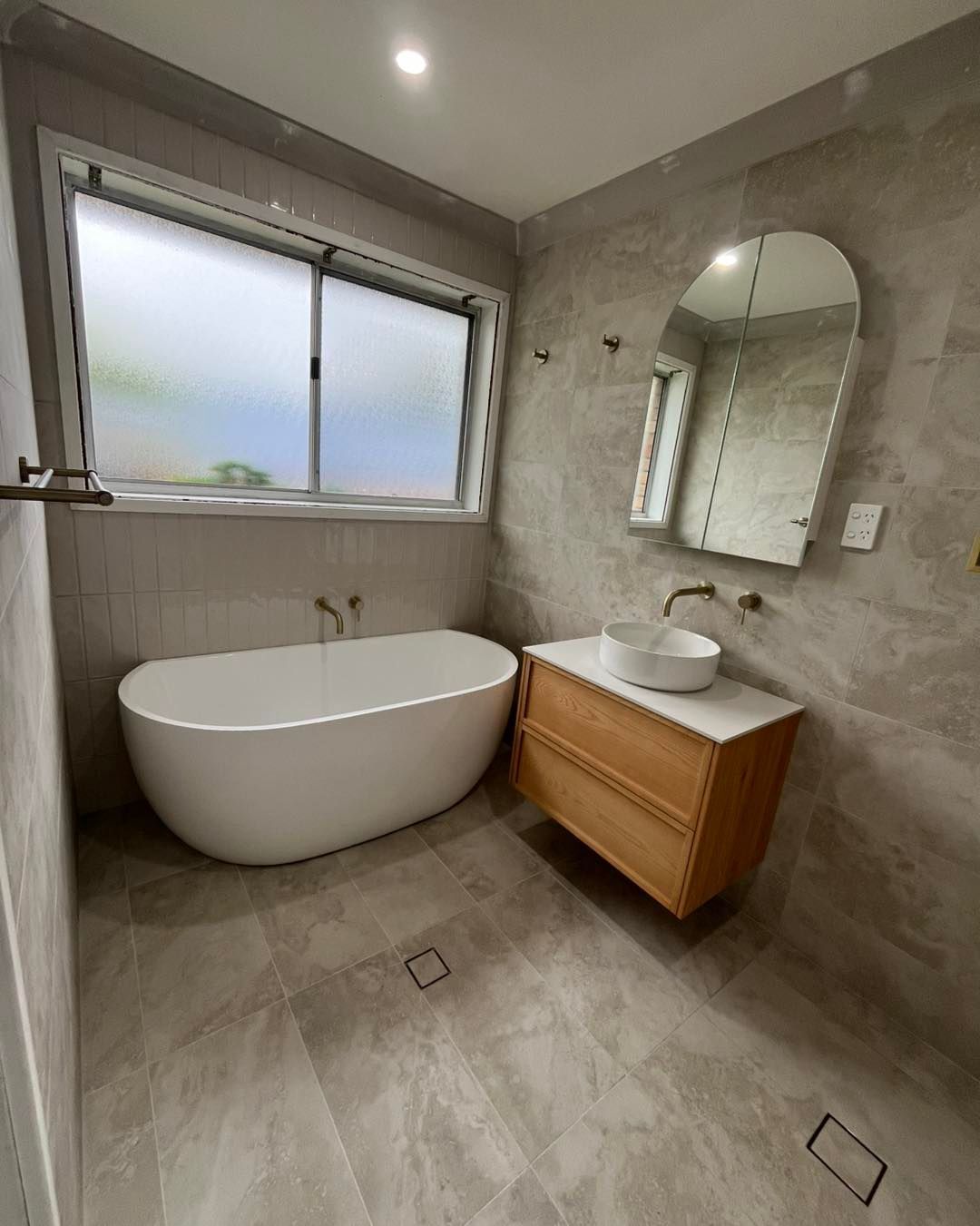 Modern Bathroom With Freestanding Tub, Wooden Vanity, and Large Arched Mirror — Cobra Plumbing & Bathrooms in Kiama, NSW