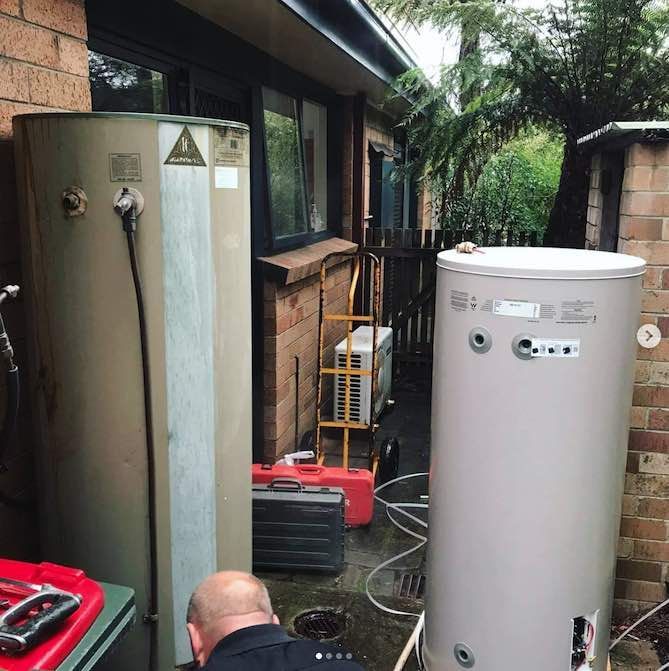 Two Water Heaters Outside a Building, With a Person Working on One — Cobra Plumbing & Bathrooms in Heathcote, NSW