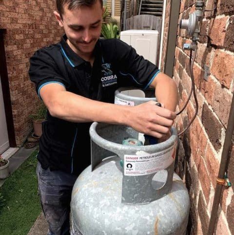 Man Connecting Gas Line to a Large Gas Cylinder Near a Brick Wall, Smiling — Cobra Plumbing & Bathrooms in Heathcote, NSW