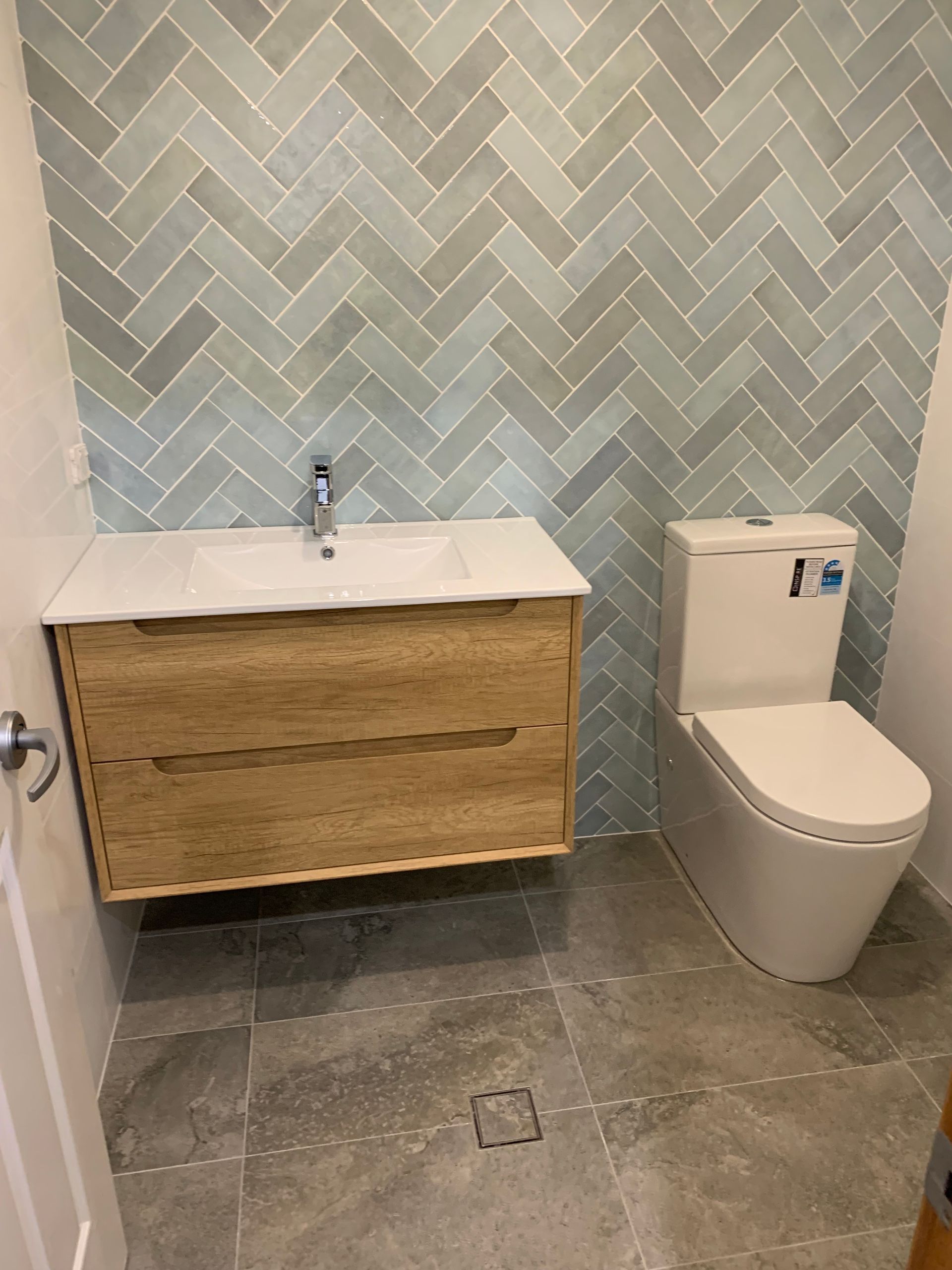 Bathroom with light wood vanity, white sink, and toilet; gray herringbone tile wall and floor — Cobra Plumbing & Bathrooms in Heathcote, NSW