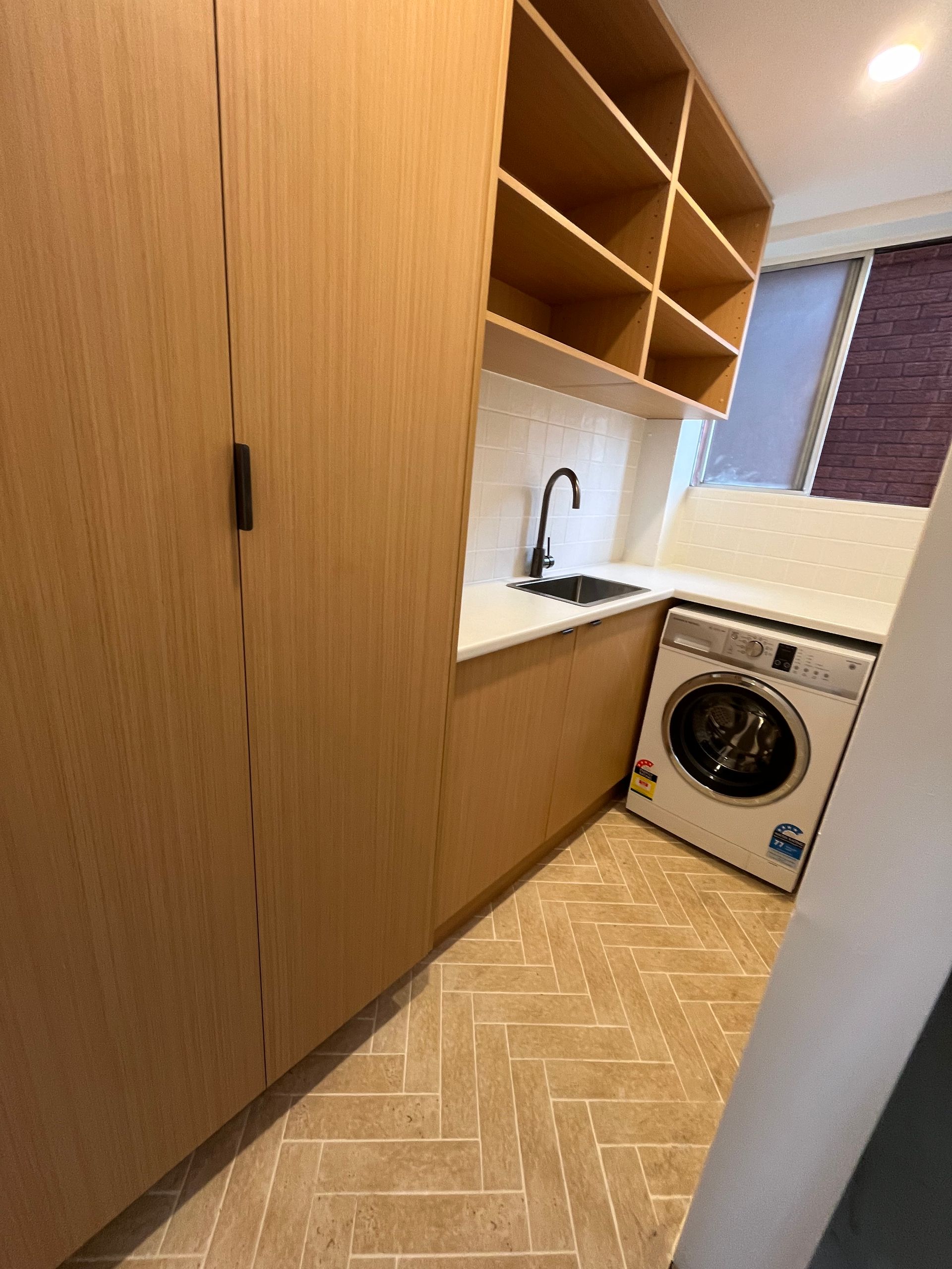 Laundry room with wooden cabinetry, sink, washing machine, and shelving. Light coloured herringbone tile flooring — Cobra Plumbing & Bathrooms in Heathcote, NSW