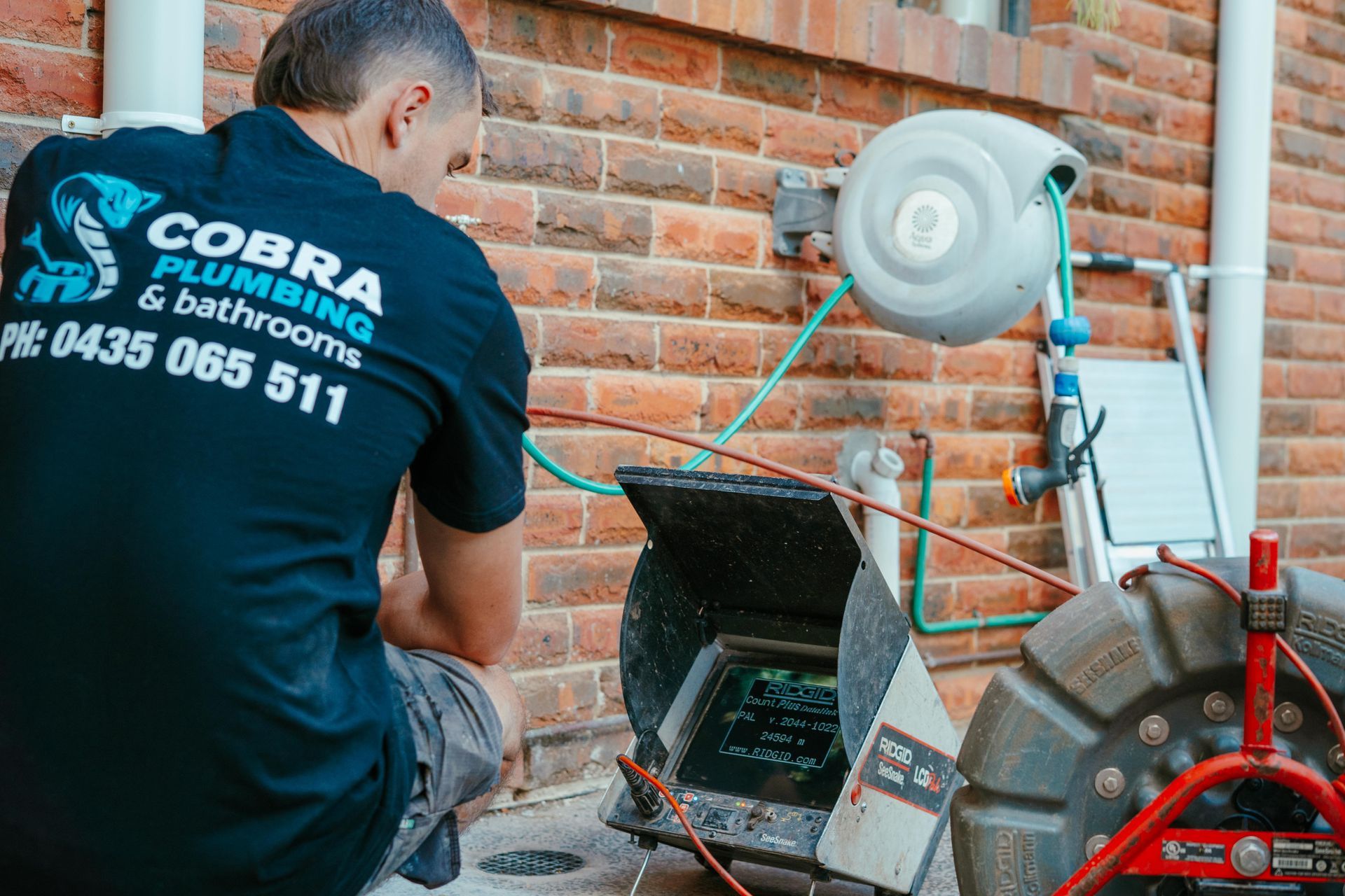 Plumber in black shirt works beside a brick wall with a drain-cleaning machine and open sewer access — Cobra Plumbing & Bathrooms in Heathcote, NSW