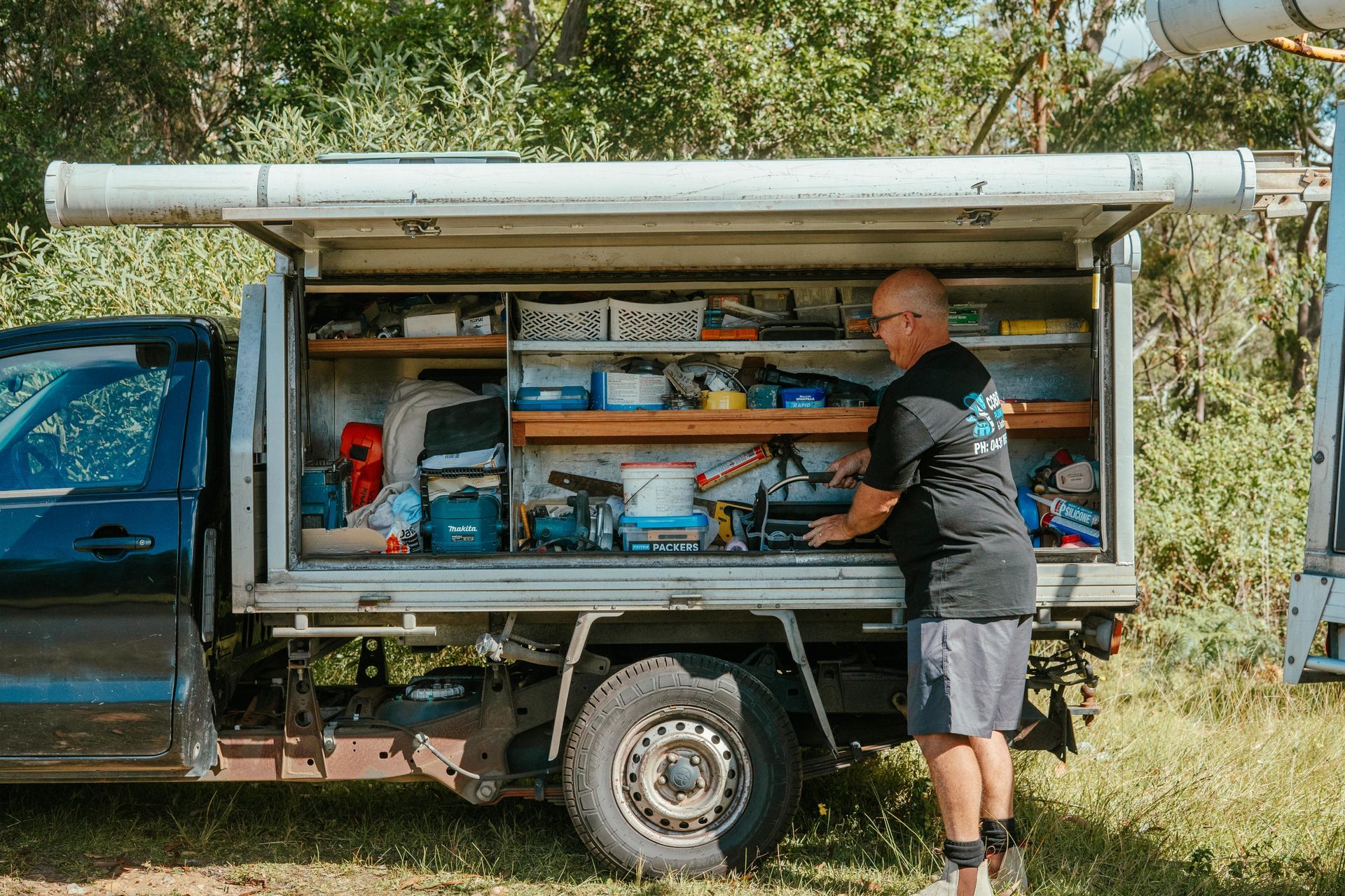Man loading tools into a blue work truck with an open side compartment outdoors — Cobra Plumbing & Bathrooms in Heathcote, NSW