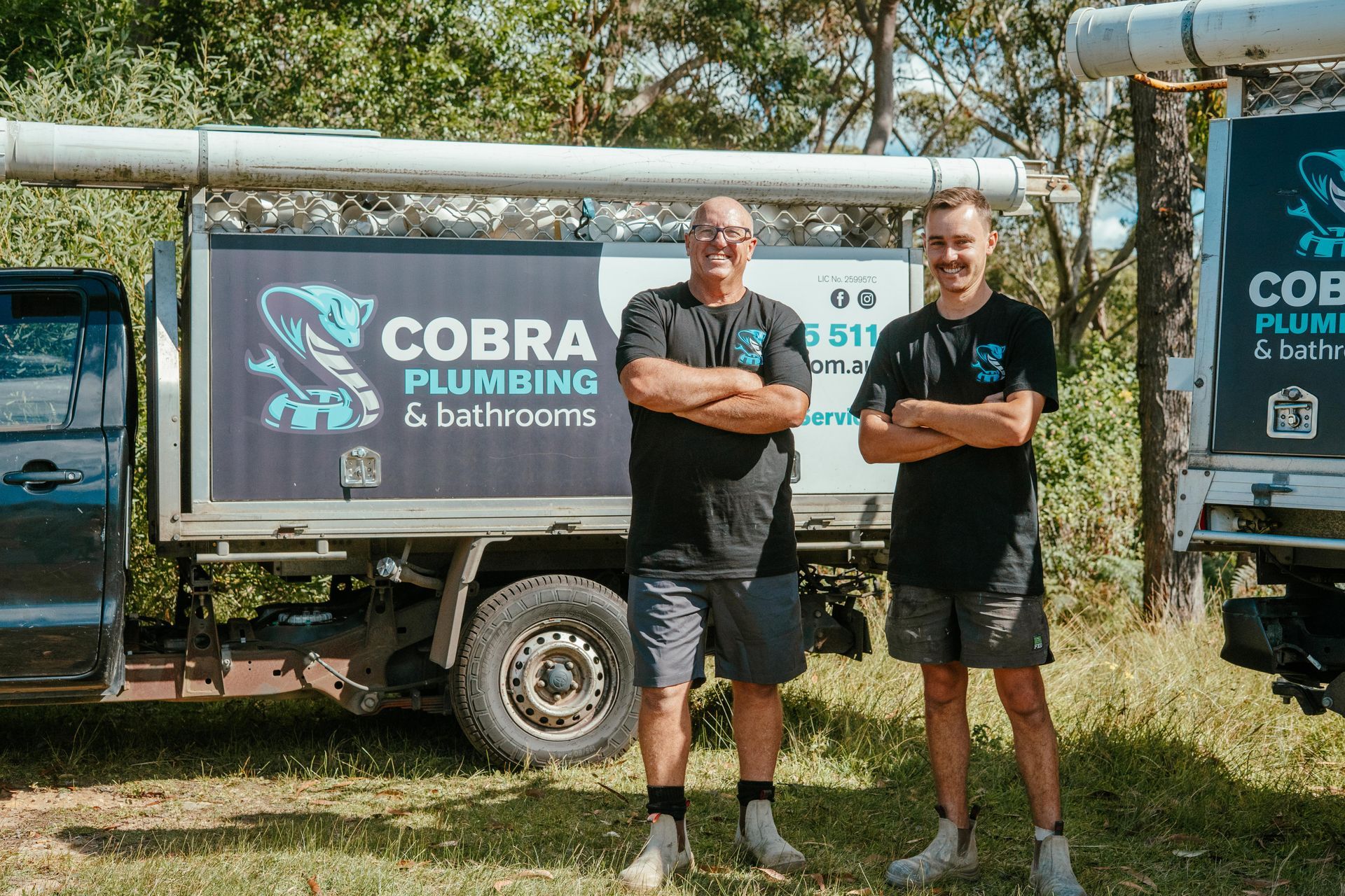 Two men standing in front of a Cobra Plumbing & Bathrooms trailer, holding a sign outdoors — Cobra Plumbing & Bathrooms in Heathcote, NSW