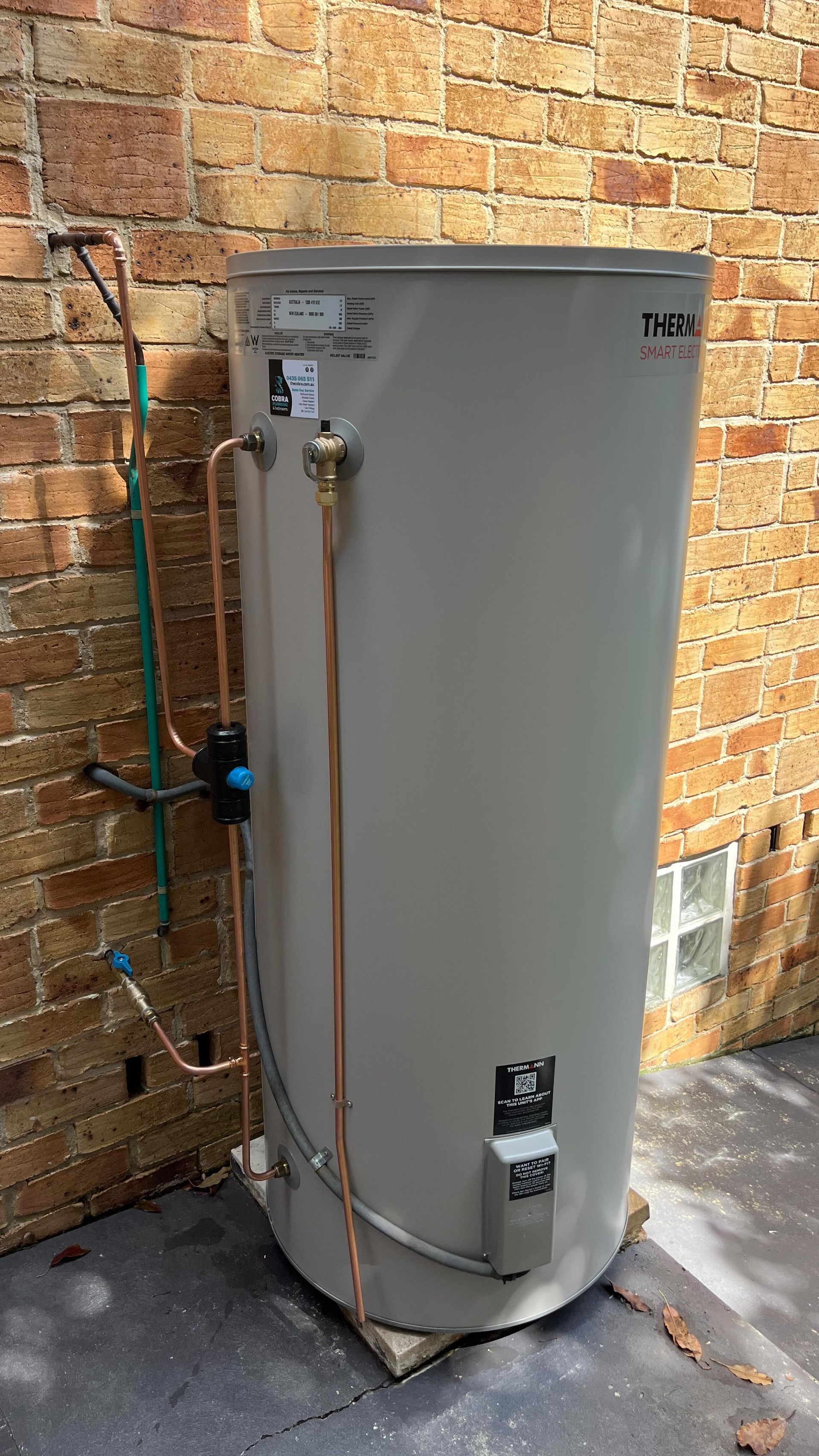 Tan water heater against a brick wall. Copper pipes and a blue valve are visible — Cobra Plumbing & Bathrooms in Heathcote, NSW
