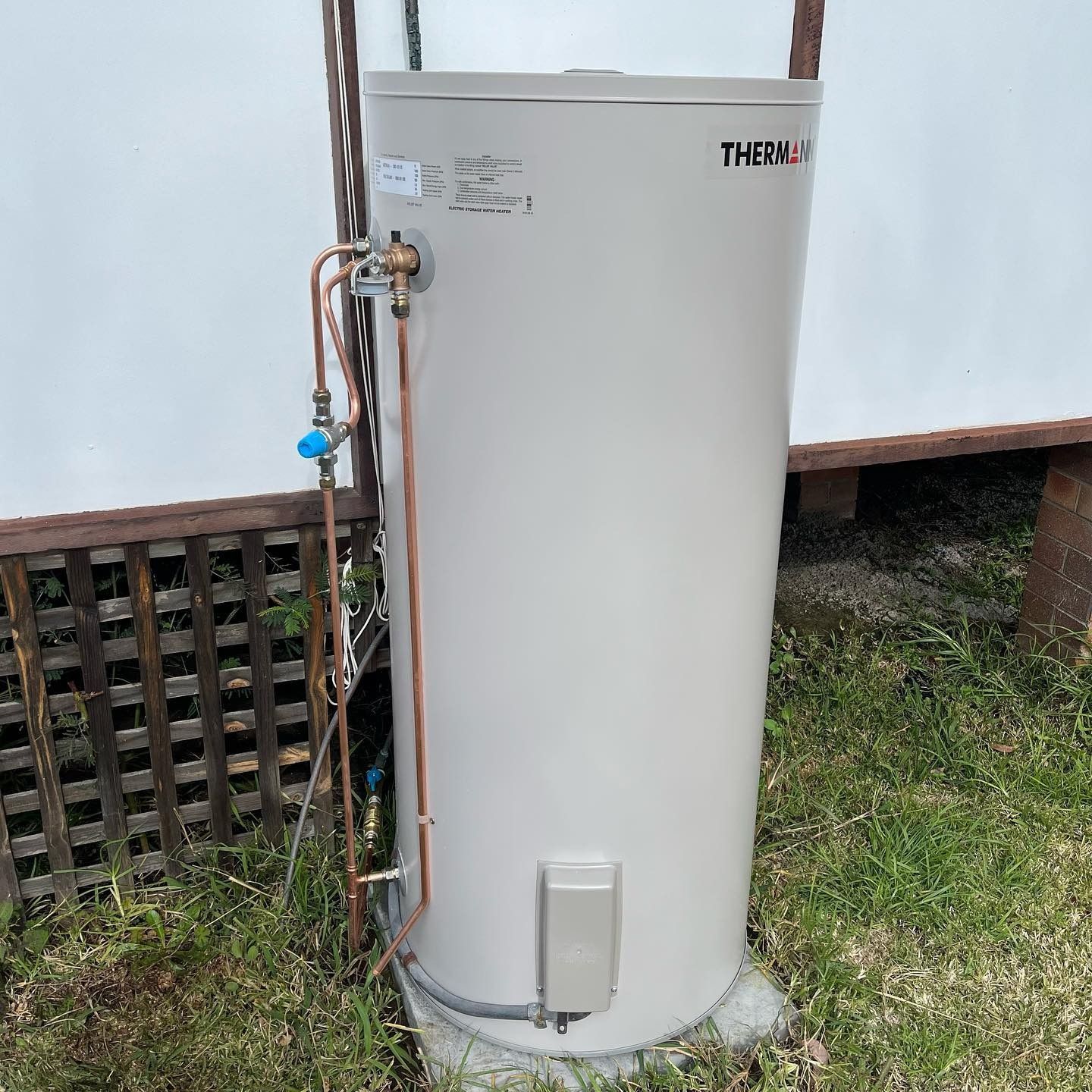 A Tall, Cylindrical, Beige Water Heater Outside Against a White Wall and Grassy Ground — Cobra Plumbing & Bathrooms in Kiama, NSW