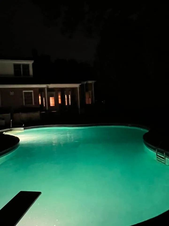 A swimming pool is lit up at night with a house in the background.