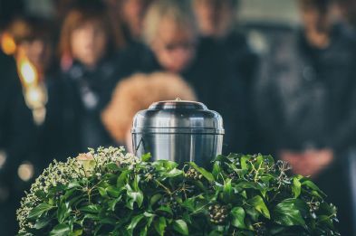 A metal funeral urn sits atop a bed of green foliage, with blurred figures in dark clothing standing in the background.