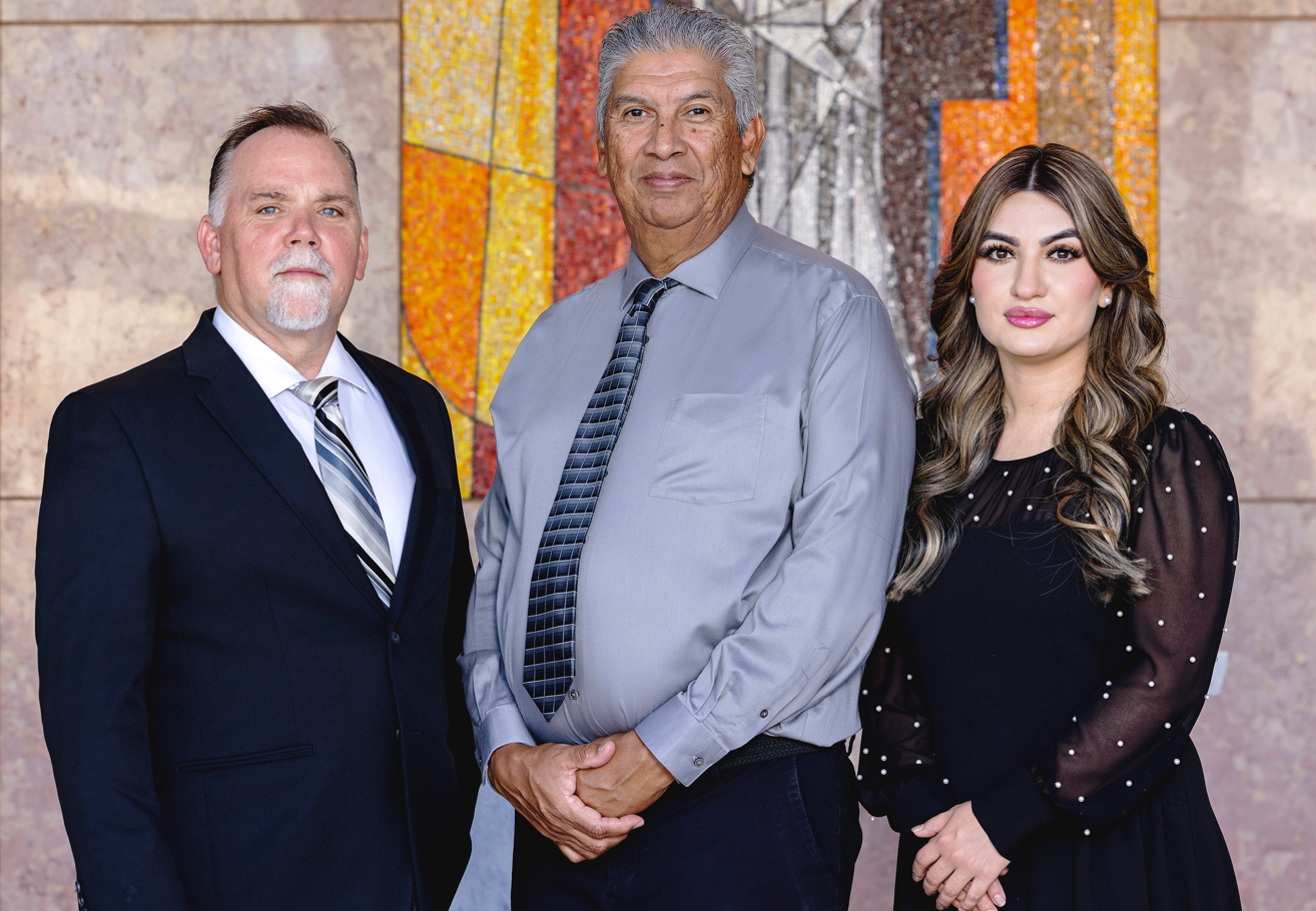 Three professionals in formal attire pose in front of a patterned wall for a studio portrait.