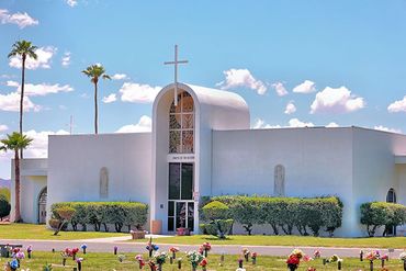 A white, modern-style mausoleum with a cross atop an arched entrance, set in a sunny cemetery.