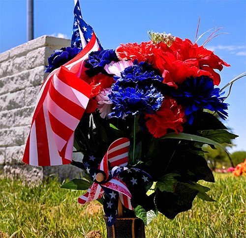 A bouquet of red, white, and blue flowers and a miniature American flag set against a cemetery backdrop.