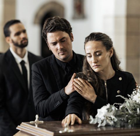Three people in black attire stand by a casket at a funeral, with two comforting each other while looking down.