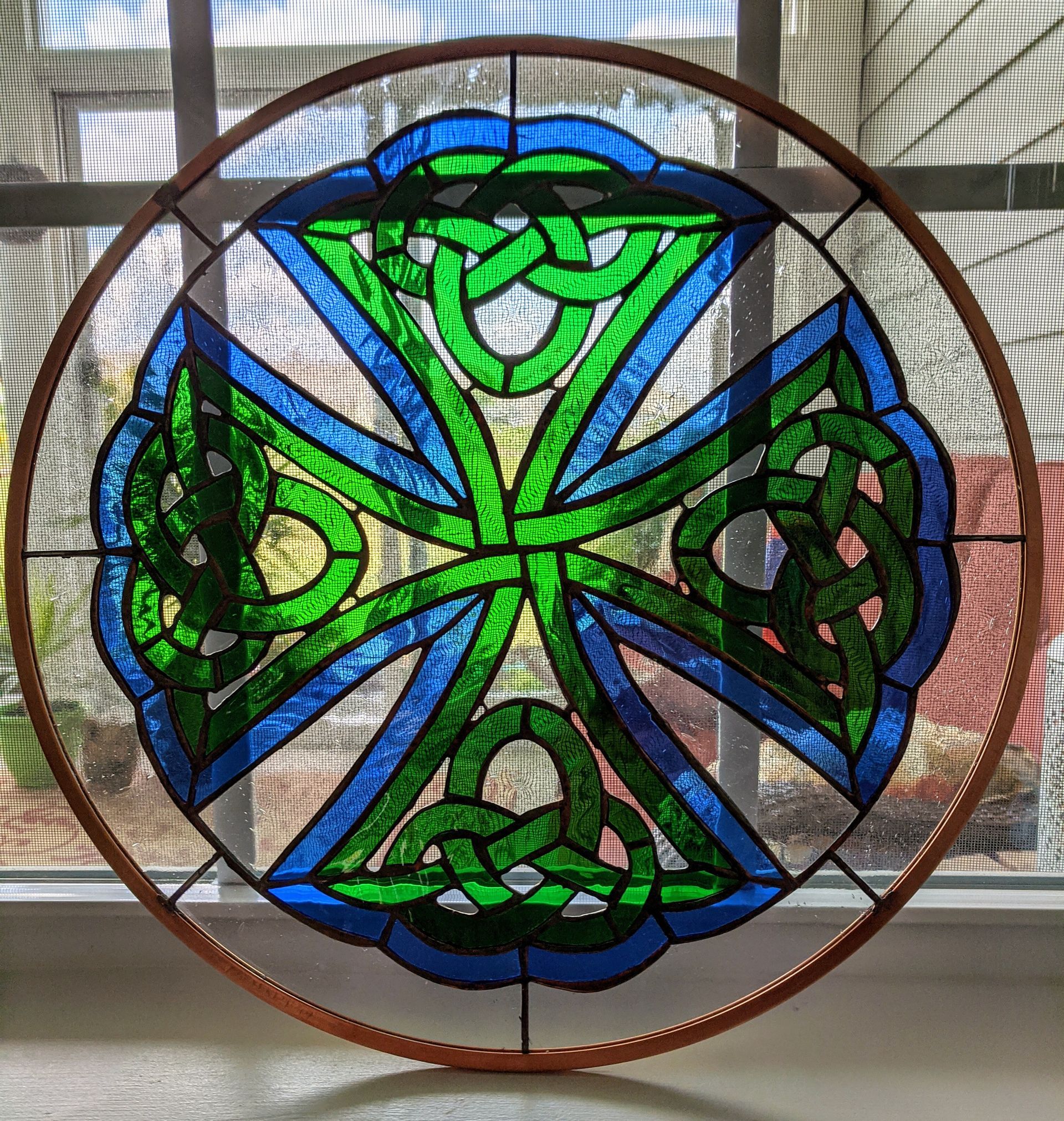Circular stained-glass-style Celtic knot ornament in green, blue, black, and white hanging in a window