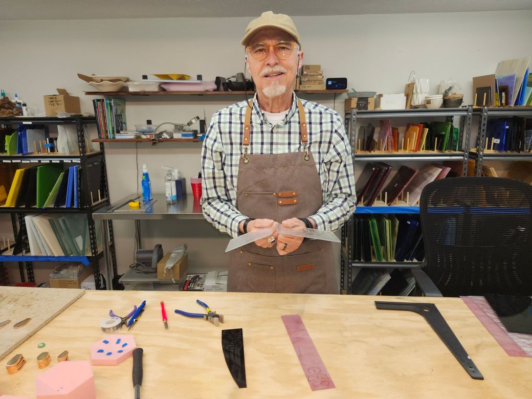 Man in a workshop holding a knife, with tools and leather pieces laid out on a worktable.