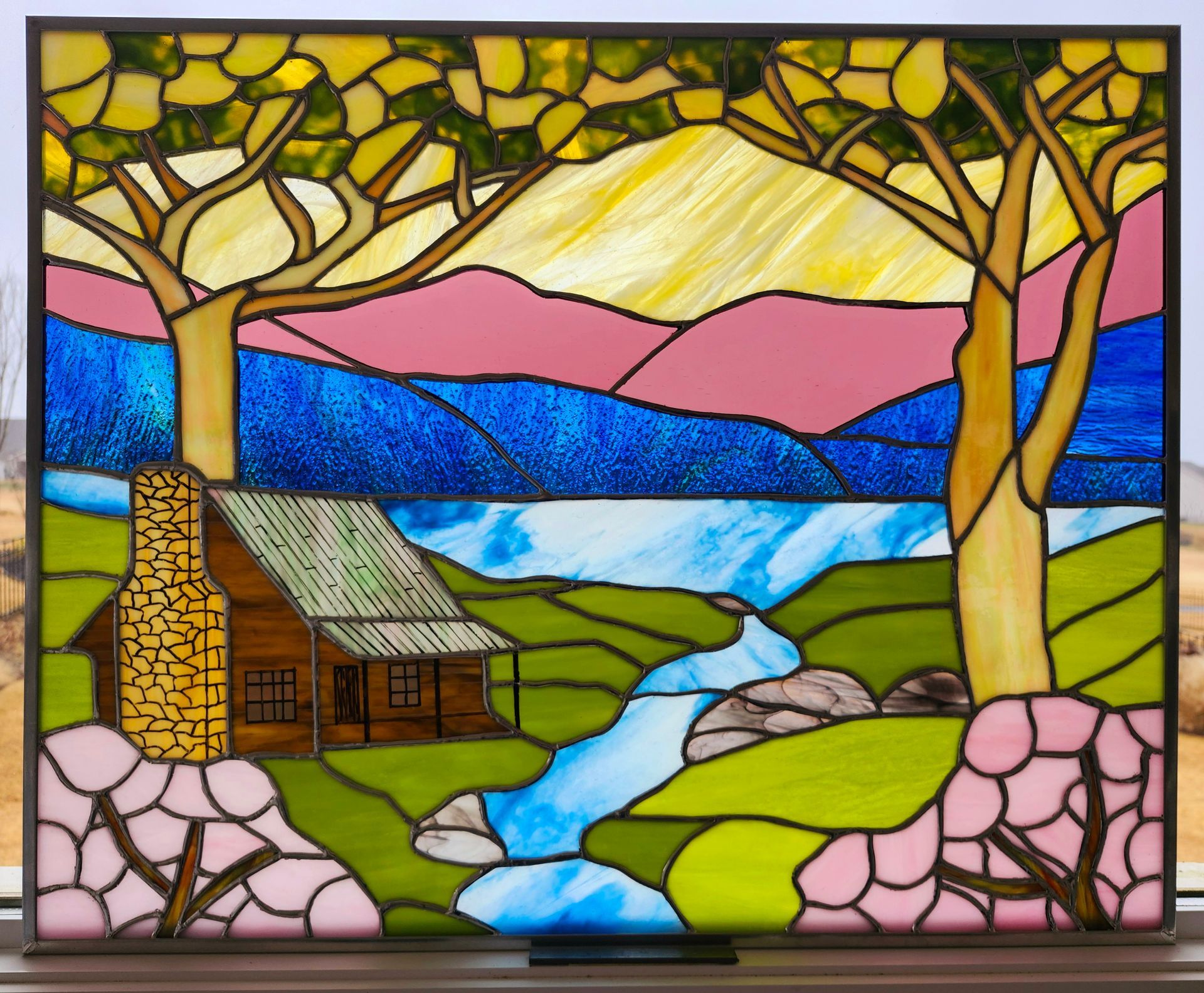 Stained-glass landscape of a cabin, river, trees, and mountains in bright blue, pink, and green.