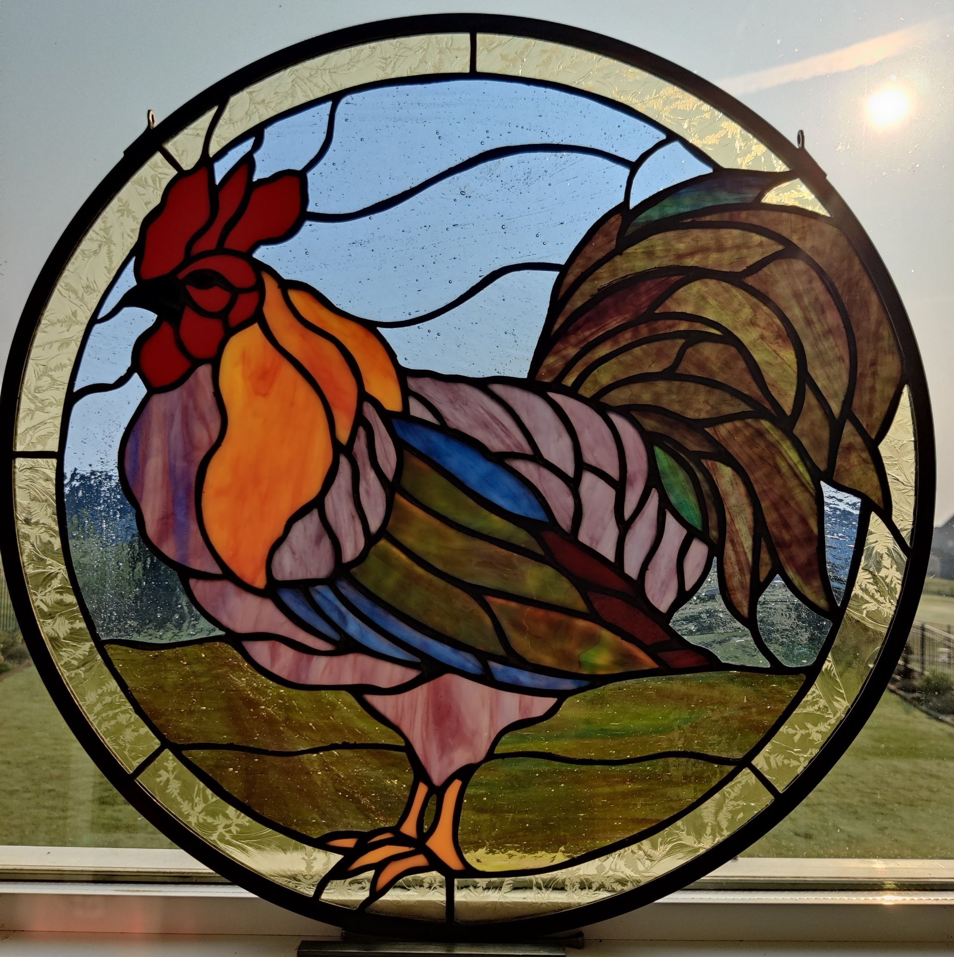 Stained glass rooster in a circular frame, with red comb, colorful feathers, and green landscape background