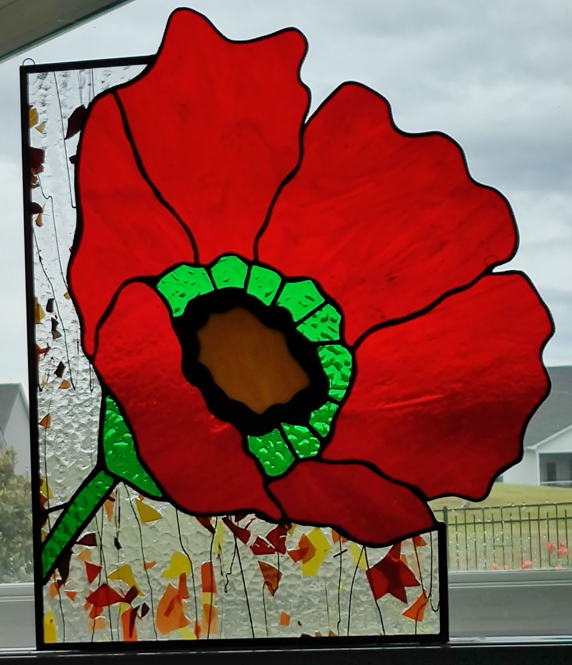 Red poppy mosaic with green center on a white panel, set against a cloudy outdoor background