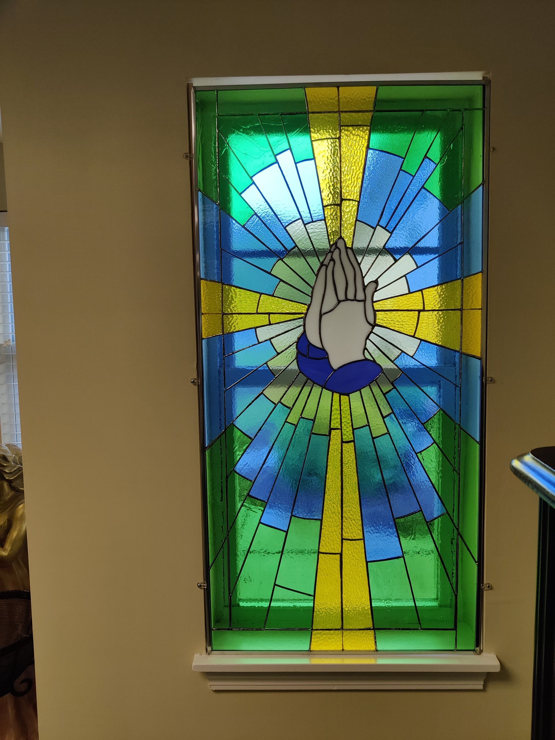 Stained-glass window with praying hands over a blue, green, and yellow sunburst design.