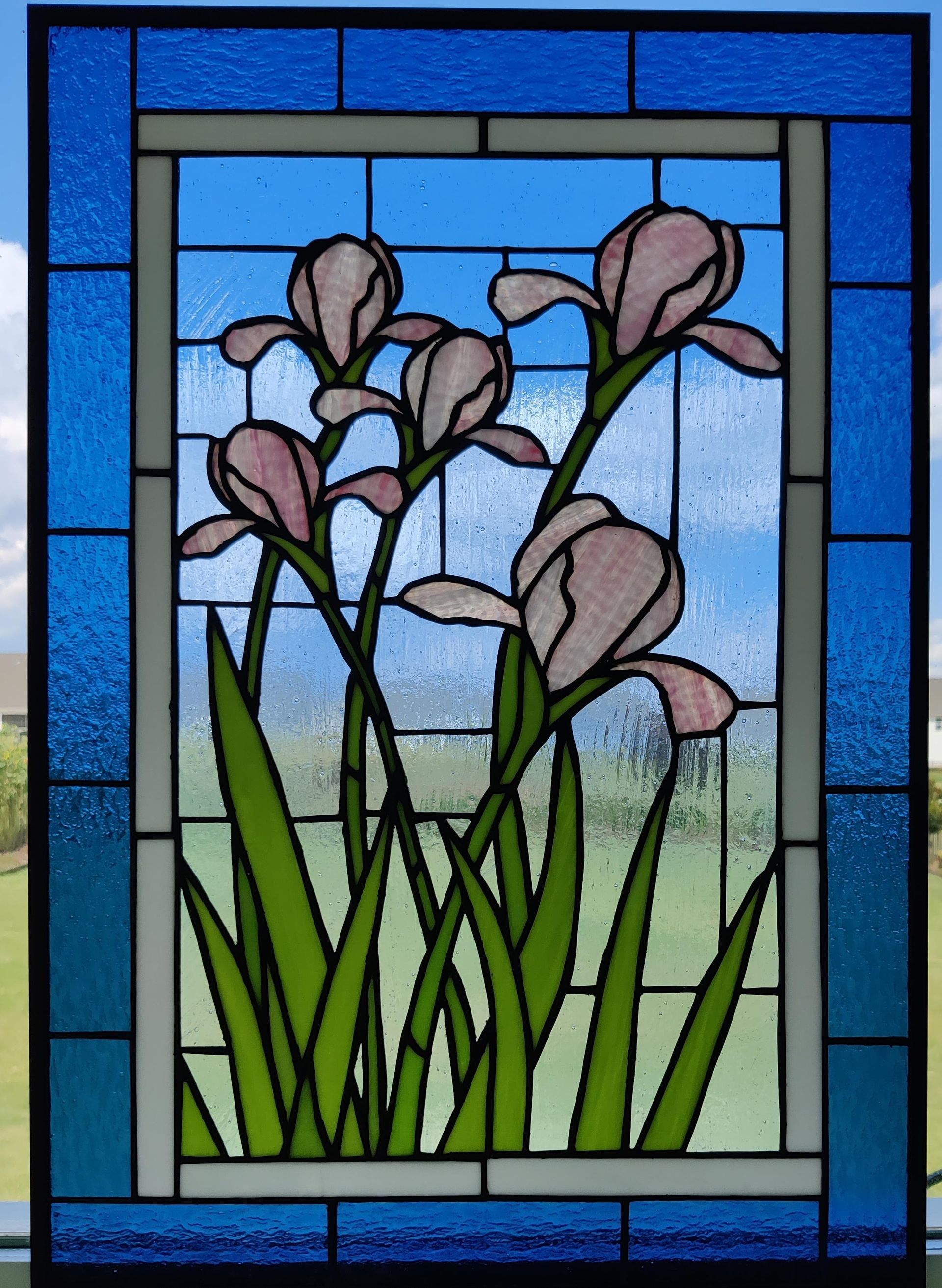 Stained glass window with purple flowers and green stems against a blue sky and grassy background