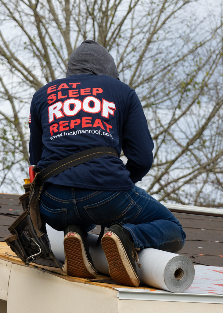A man is kneeling on a roof wearing a shirt that says `` eat sleep roof repeat ''.