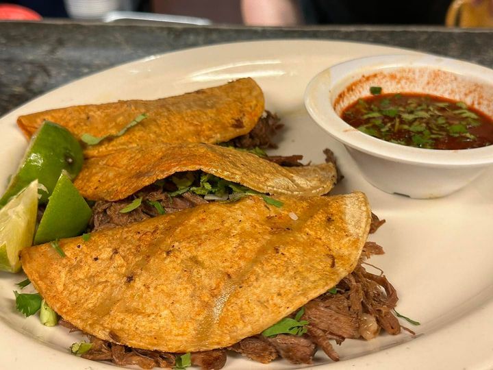 Three beef tacos with lime wedges and a side of red dipping sauce on a white plate.