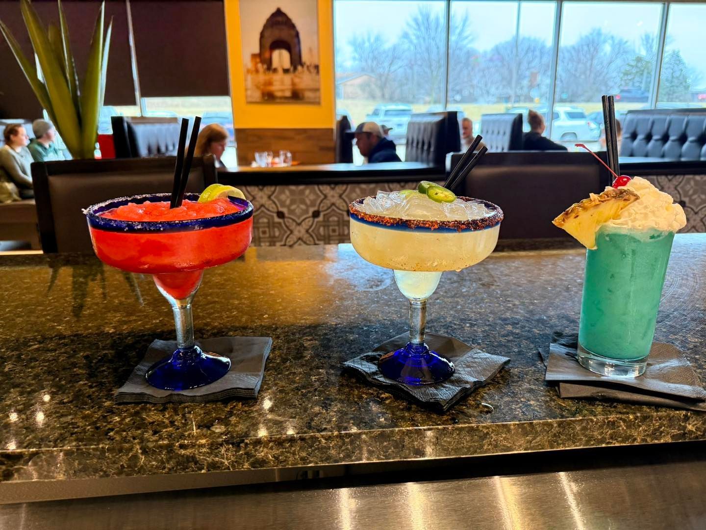 Three colorful cocktails served in a row on a granite bar top in a restaurant setting.