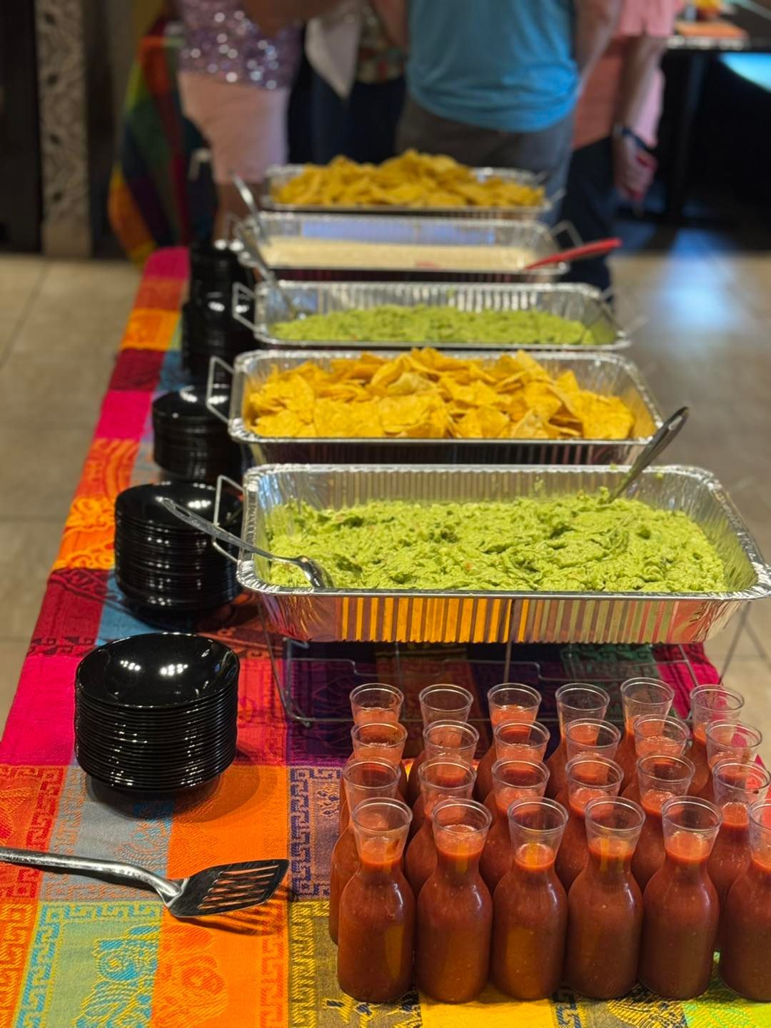 A buffet table with aluminum trays of chips, guacamole, and salsa, next to stacks of plates and small bottles of red sauce.