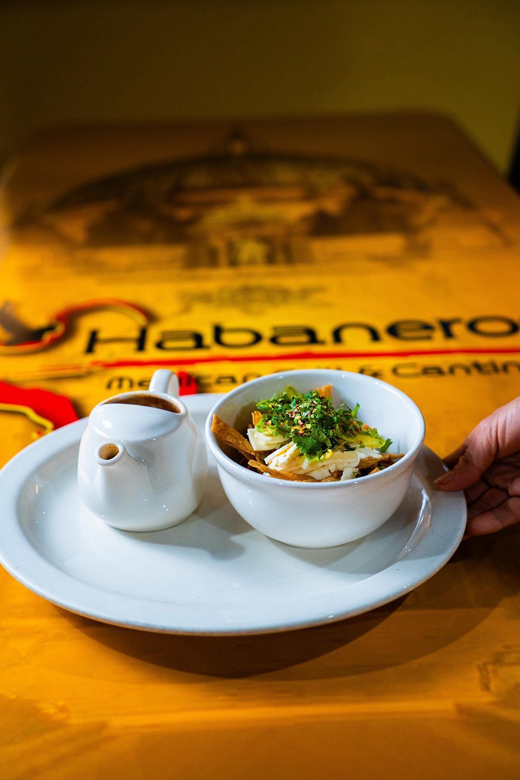 A bowl of chilaquiles with a side of sauce on a white plate held over a Habanero restaurant menu.