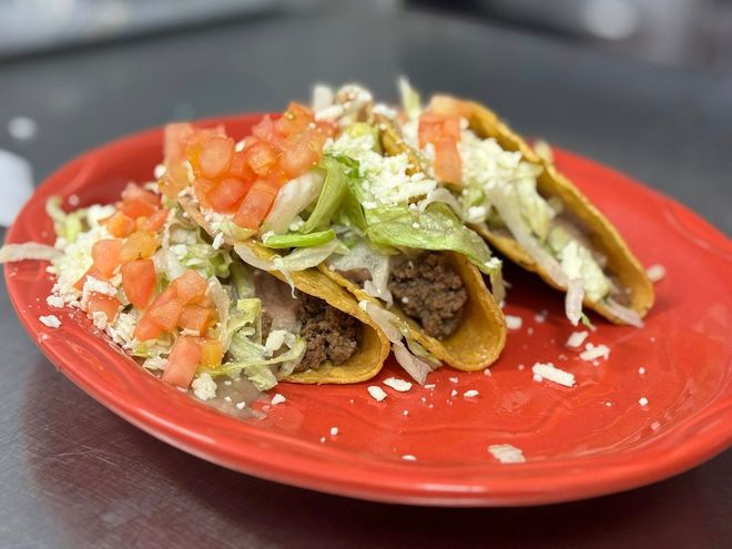 Three crispy tacos topped with lettuce, diced tomatoes, and crumbled cheese on a red plate.