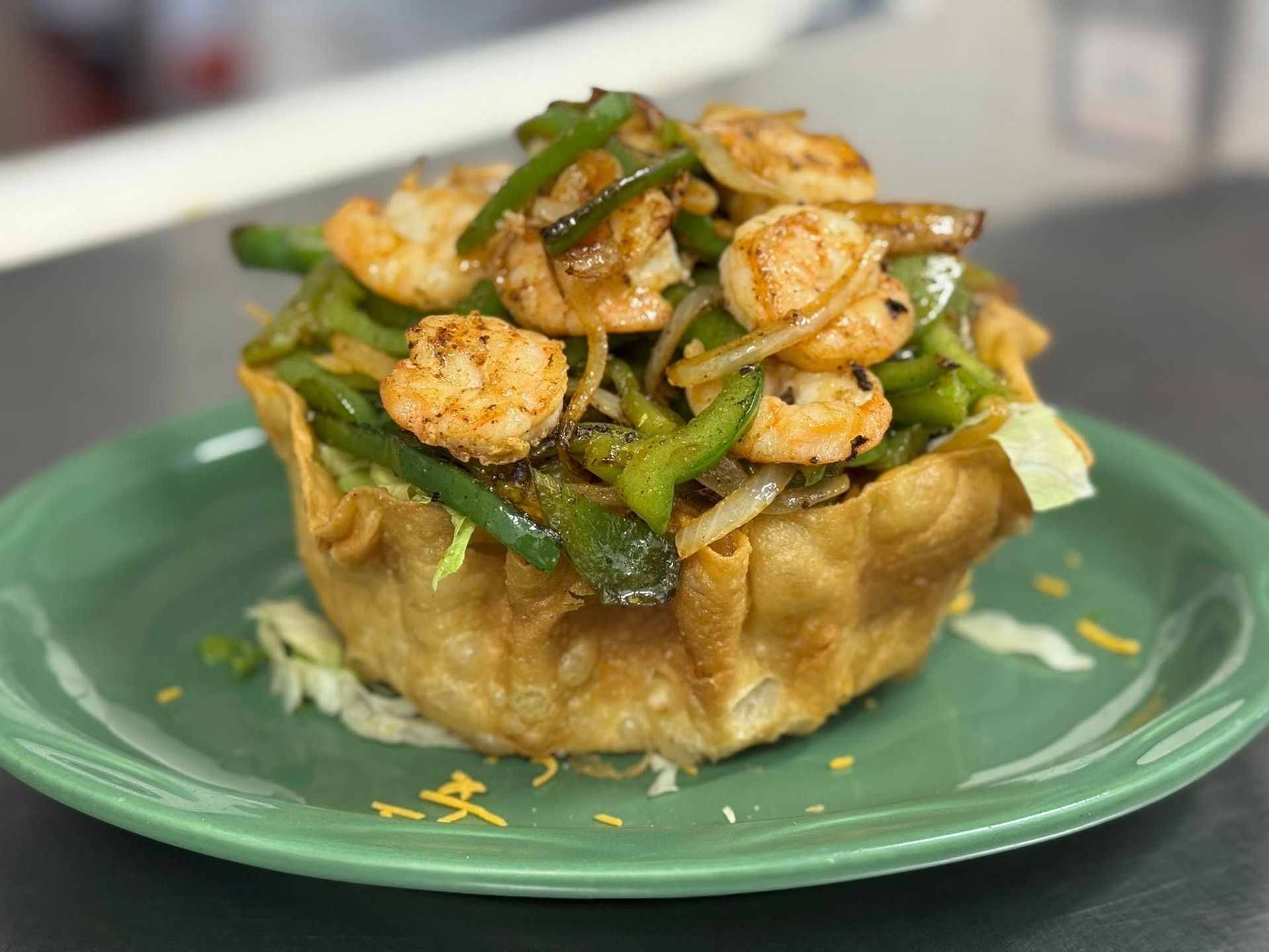 Shrimp fajitas with green peppers and onions served in a crispy, golden fried tortilla bowl on a green plate.