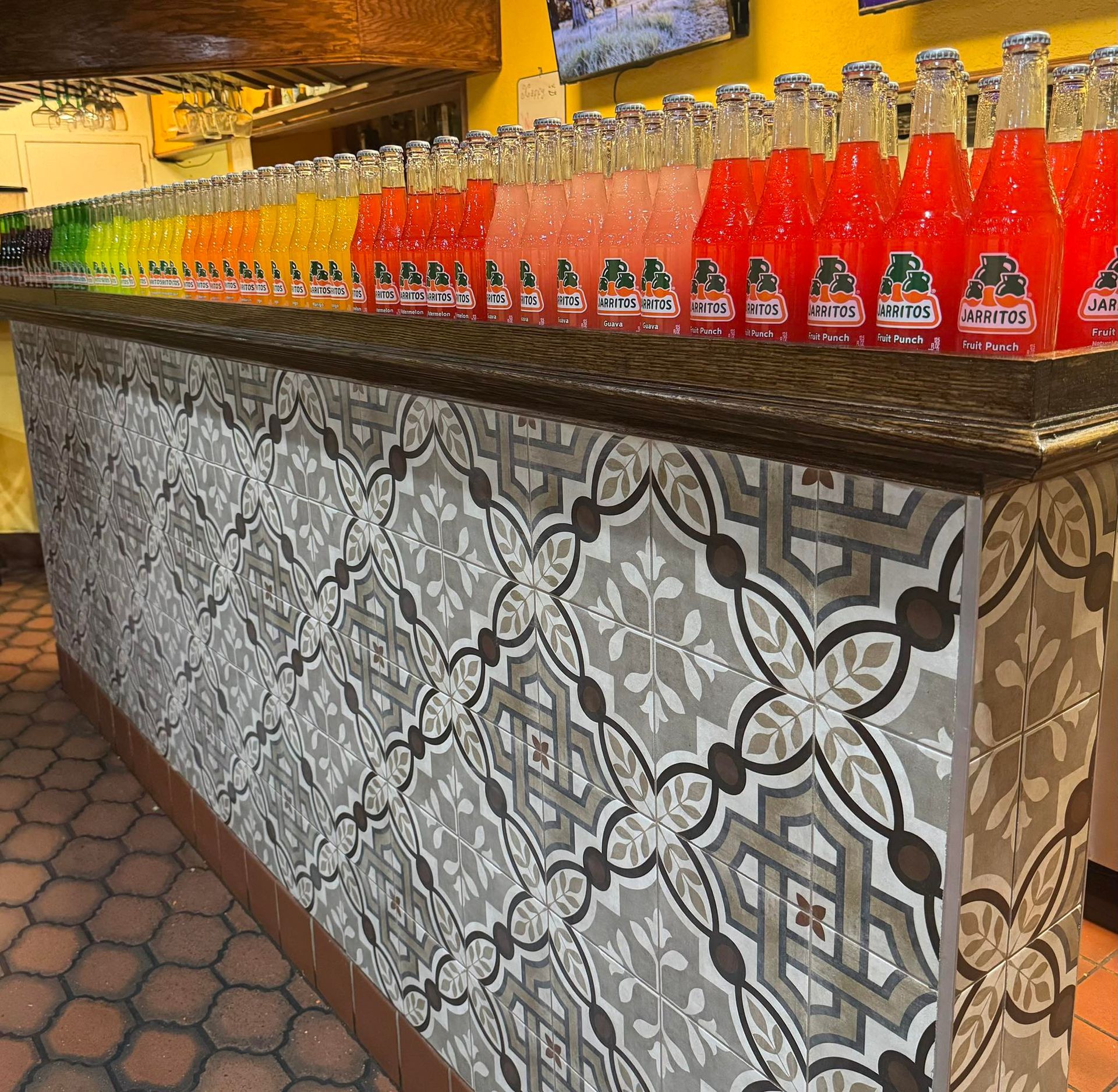 A bar counter with patterned grey and white tiles, topped with a long, colorful row of Jarritos soda bottles.