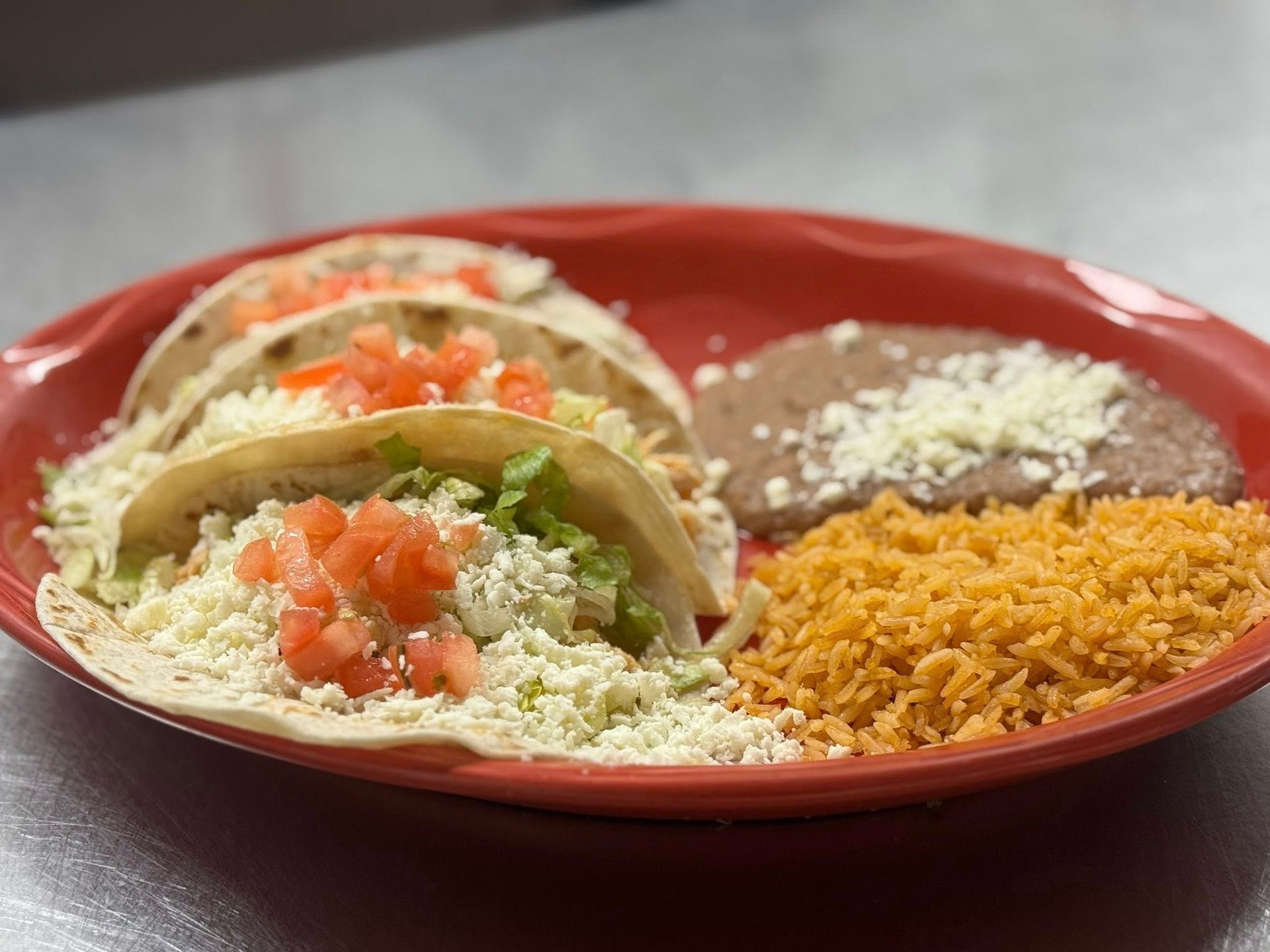 A red plate holding three soft tacos topped with cheese and diced tomatoes, served with refried beans and Mexican rice.