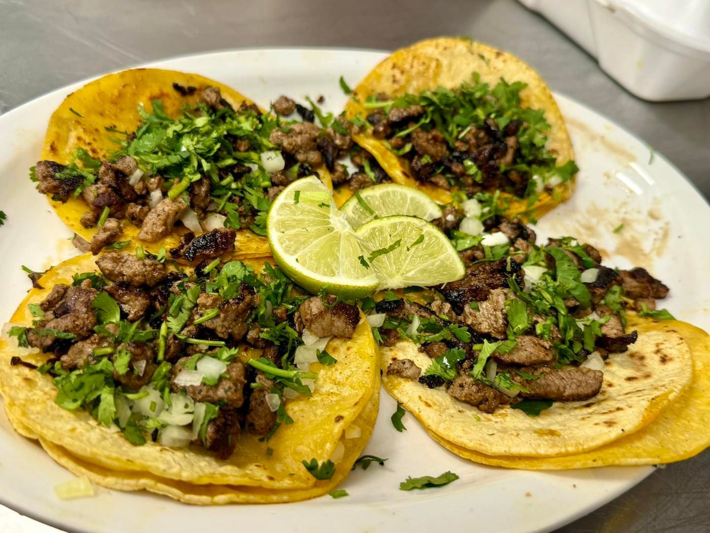 Four street tacos with steak, cilantro, and onions on yellow corn tortillas, served with two lime wedges on a white plate.