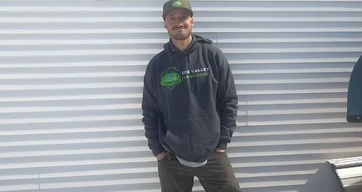 Man in green cap and hoodie leans against a white-striped wall, hands in pockets.
