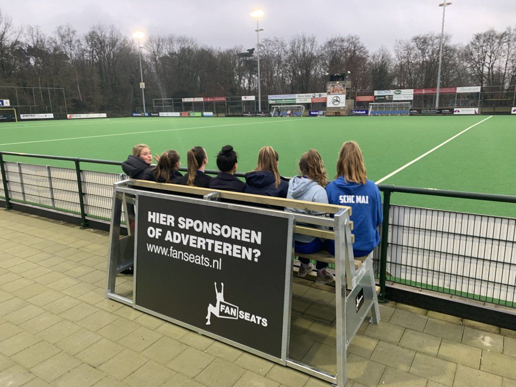 Een groep mensen zit op een bankje met uitzicht op een hockeyveld. Op het bankje staat de tekst 